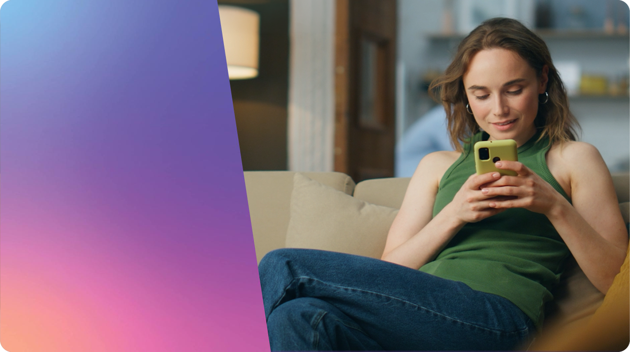 A woman with wavy brown hair sits relaxed on a beige couch in a softly lit, modern living room, smiling down at a green smartphone she's holding with both hands. She wears a green sleeveless top and blue jeans, with hoop earrings, a floor lamp and shelving visible in the blurred background. The left third of the frame is a smooth diagonal gradient blending sky blue, violet, and coral-pink — likely reserved for a title/logo overlay.