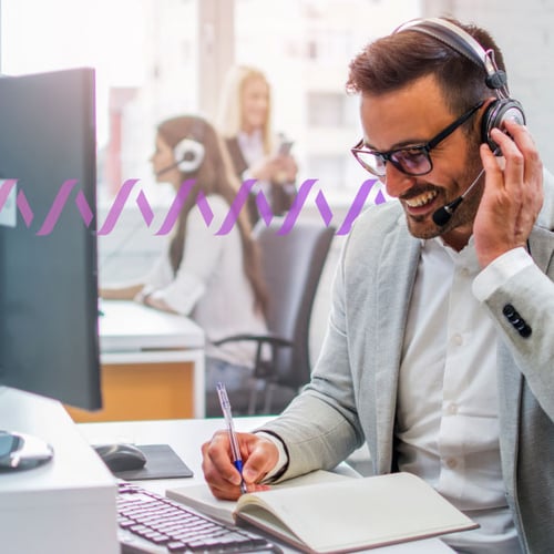 Photo of a smiling agent in an office setting talking to a customer through his phone headset while taking notes. In the background, a series of small purple waves runs across the frame..