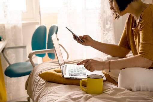 woman doing remote work from bed with laptop and mobile phone
