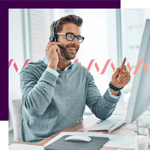 Photo of a smiling contact center agent sitting in front of his computer and talking to a customer through his phone headset. In the background, a small gradient-pink waveform runs across the frame.