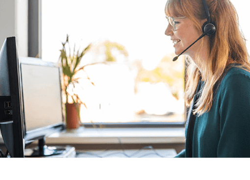 UC Cloud Call Center Article Header Photo of a smiling female call center agent talking to a customer on her headset as she types notes on her computer.