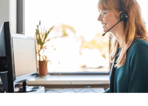 Photo of a smiling female call center agent talking to a customer on her headset as she types notes on her computer.