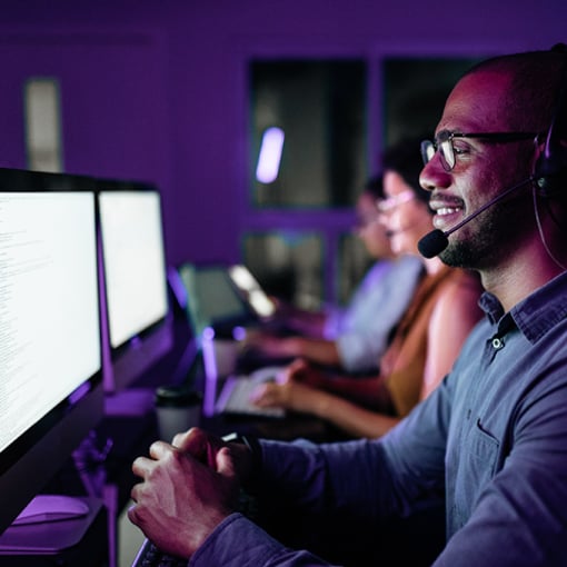 VCC Call Center Software Article Thumbnail Photo of a call center; we see a side view of a row of working agents in a darkened room, their faces illuminated by the monitor screens in front of them.