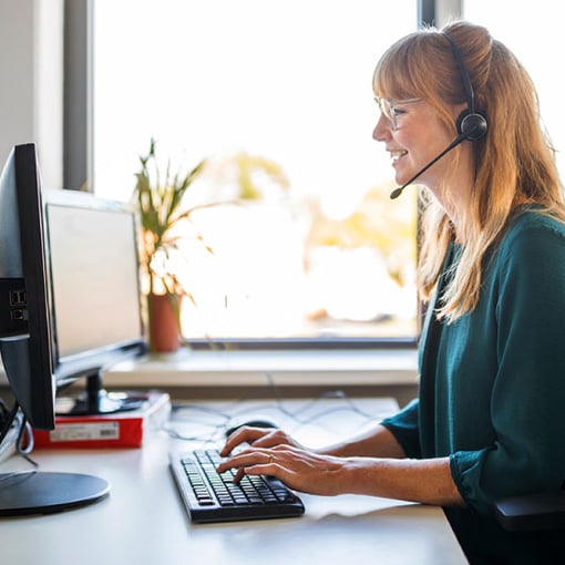 UC Cloud Call Center Article Thumbnail Photo of a smiling female call center agent talking to a customer on her headset as she types notes on her computer.