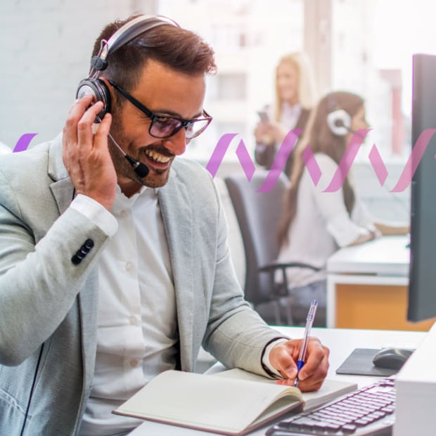 UC Call Center QA Blog Header Photo of a smiling male call center agent talking to a customer through his headset and taking notes