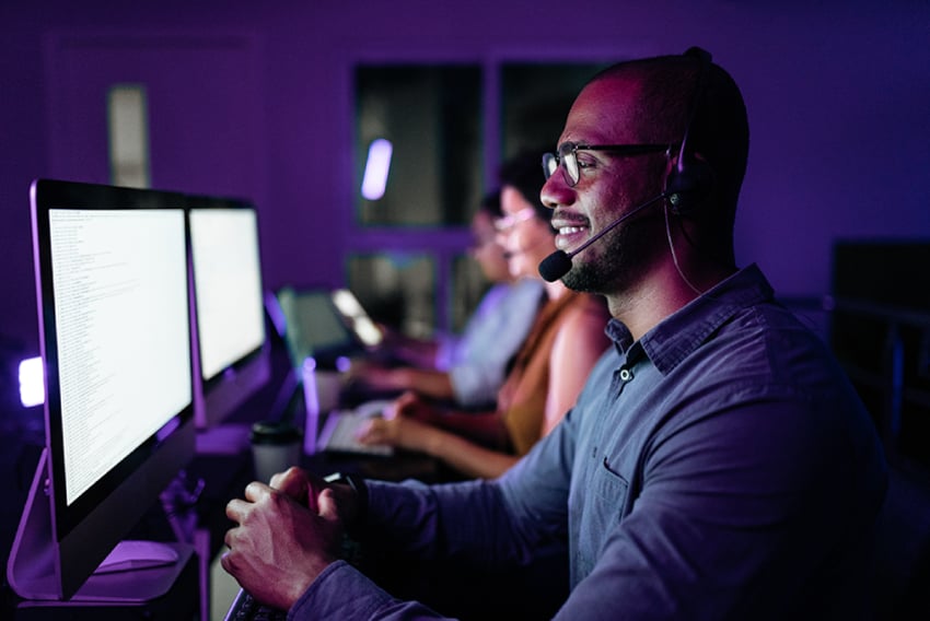 Photo of a call center; we see a side view of a row of working agents in a darkened room, their faces illuminated by the monitor screens in front of them.