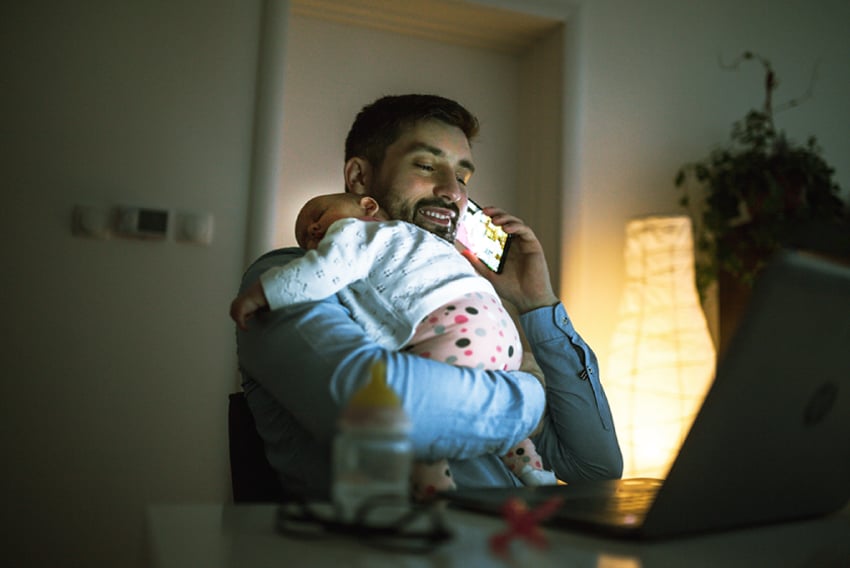 UC Choosing VoIP Provider Article Header Photo of a business man at home, talking on his cellphone. He is holding a sleeping infant over his shoulder.