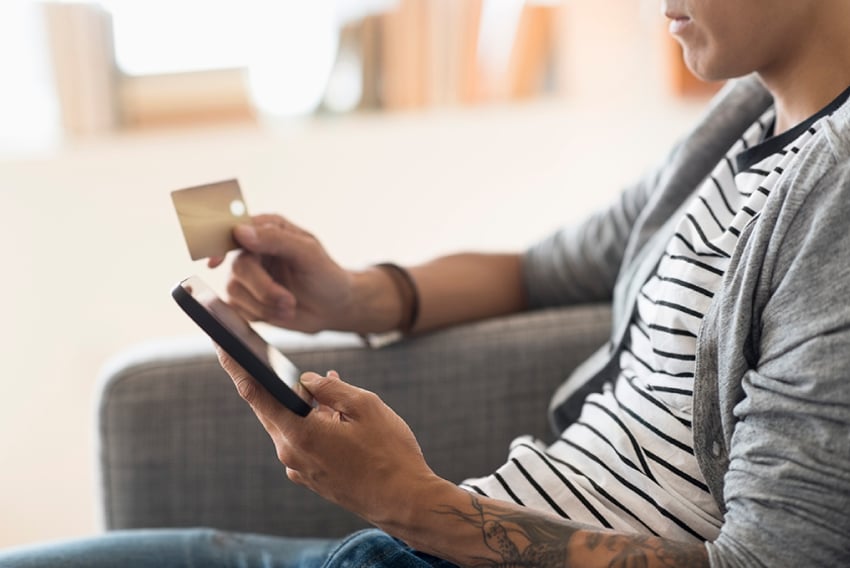 Photo of a man sitting in a chair. In one hand he is holding his cellphone; in the other he is holding a credit card as he makes a purchase with his phone. 