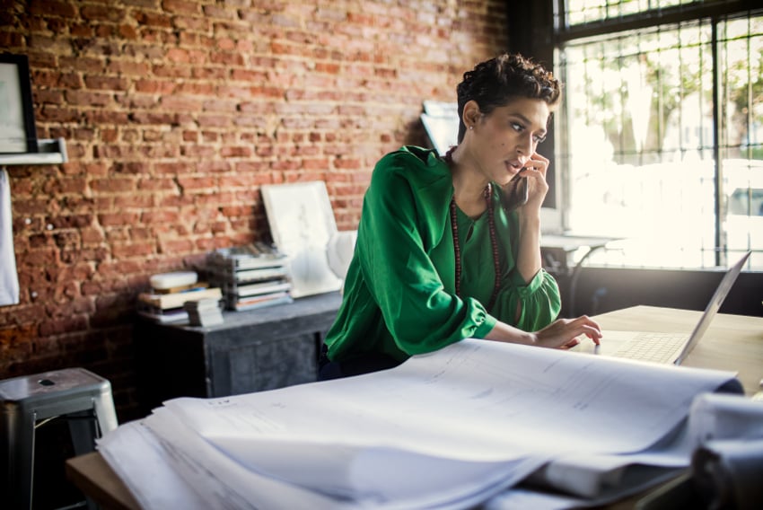 Stock photo of woman, a small business owner who is on her cell phone and typing notes into her laptop.