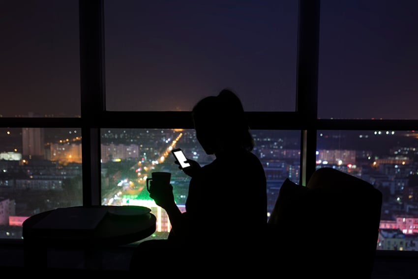 UC Use VoIP Without Internet Article Header Photo of a woman in an office at night. The lights are out, and we can see her in the glow of her cellphone and the lights of a city outside her window.