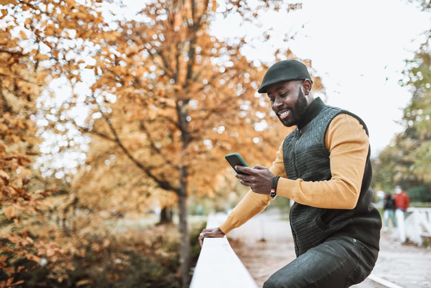 Photo of a smiling man standing outdoors. He is looking at something on his phone.