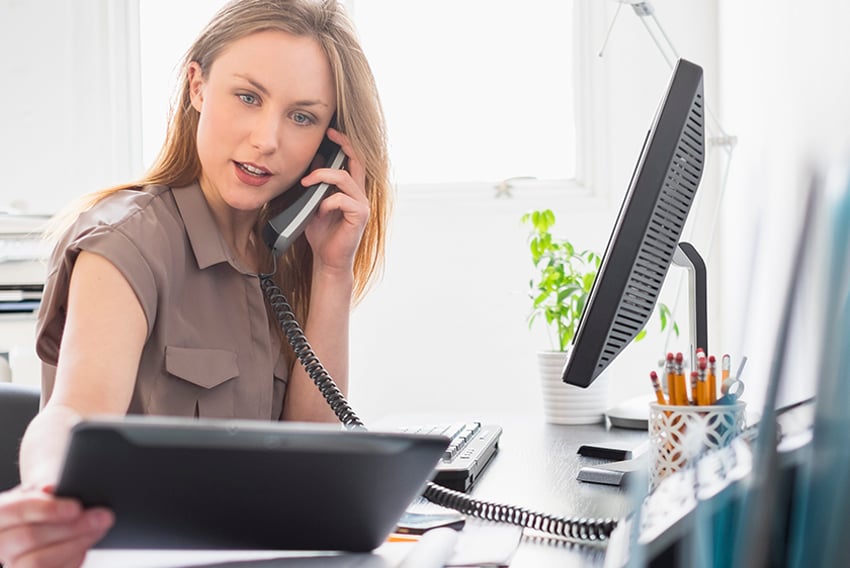 Woman sitting at a desk, on the phone, looking at a tablet. This conveys VoIP phone service. Woman sitting at a desk, on the phone, looking at a tablet. This conveys VoIP phone service.