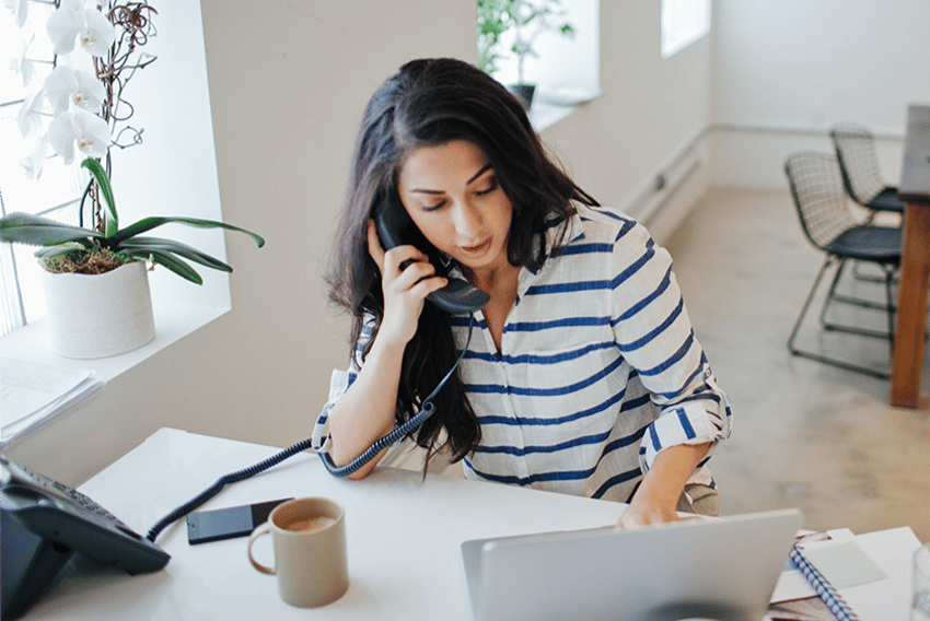 UC VoIP vs. Landline Header photo of a woman sitting at a desk, working on her laptop. On the desk is a traditional landline office phone, which she is holding up to her ear, and a cellphone.