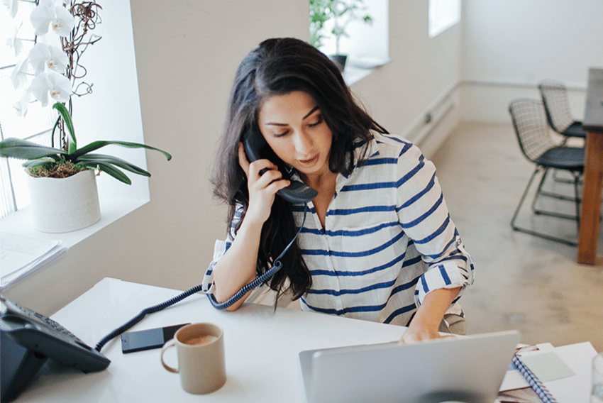 photo of a woman sitting at a desk, working on her laptop. On the desk is a traditional landline office phone, which she is holding up to her ear, and a cellphone.