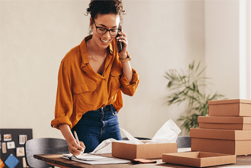 What Is VoIP Phone System Header Photo of a smiling woman standing at her busy, cluttered desk in what appears to be a home office. She is holding her cell phone, listening and taking notes.