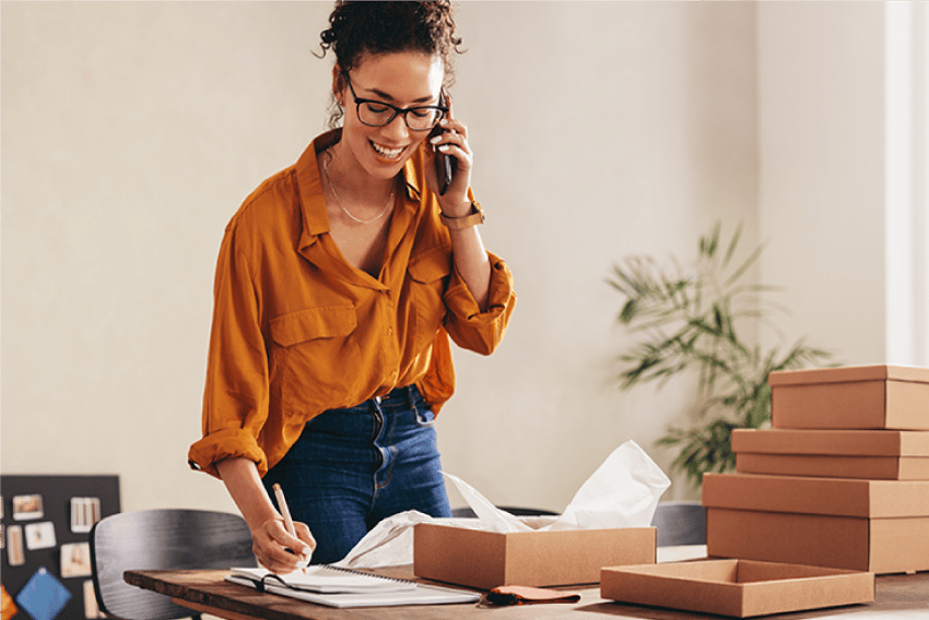 Photo of a smiling woman standing at her busy, cluttered desk in what appears to be a home office. She is holding her cell phone, listening and taking notes.