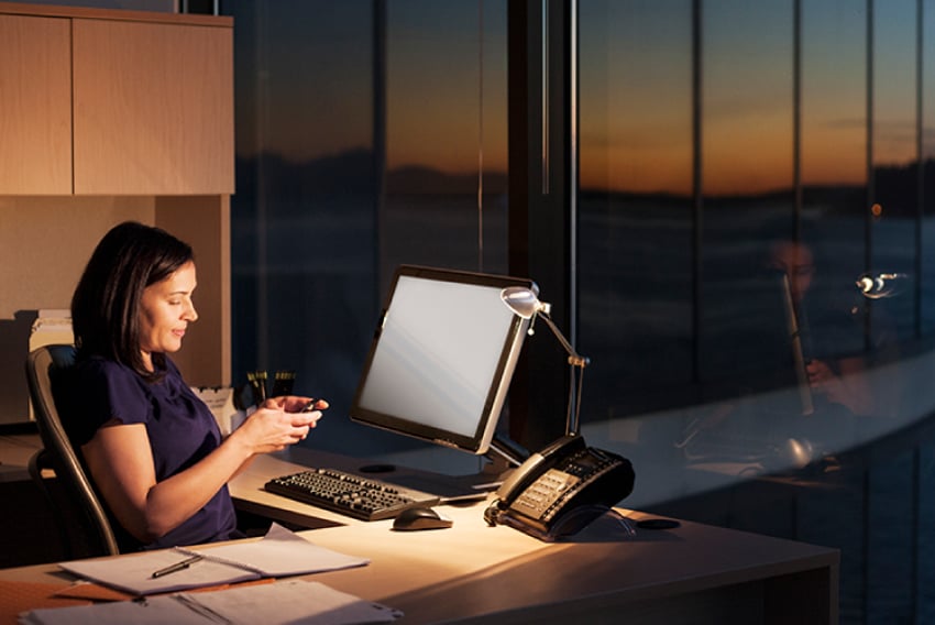 Photo of a business woman sitting in her office in the evening. A desk lamp illuminates her computer, a desktop phone, and several papers. She is holding her cellphone, reading something on the screen.