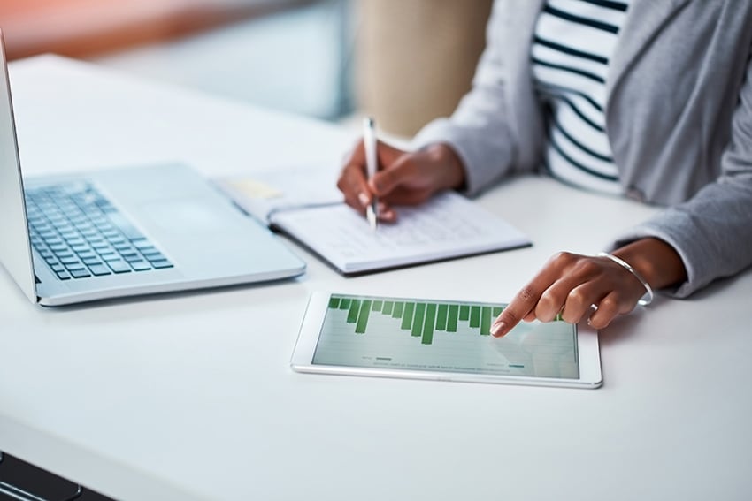 Person writing down financial info from chart and laptop in front of her