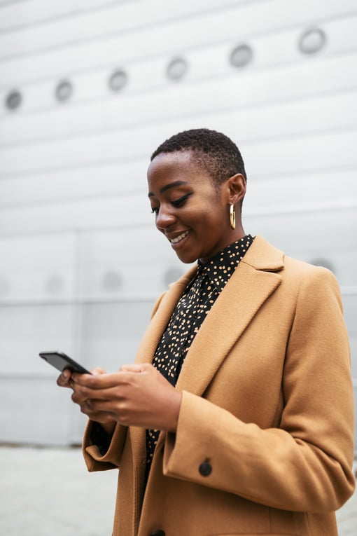 Cheerful African American female in coat messaging smartphone while standing by minimalist building on street of modern city