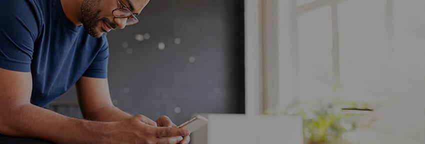 man wearing glasses looking down at phone opposite a window